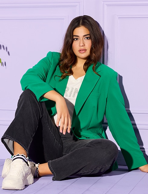 The model poses sitting in a green blazer and dark jeans 
