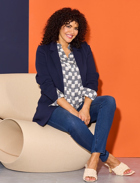 The model poses seated and wears a blouse, blazer and jeans