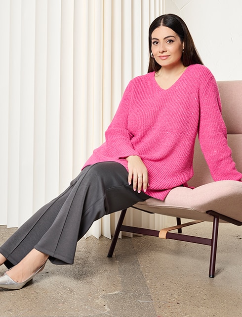 The model is wearing a pink jumper and grey trousers.
