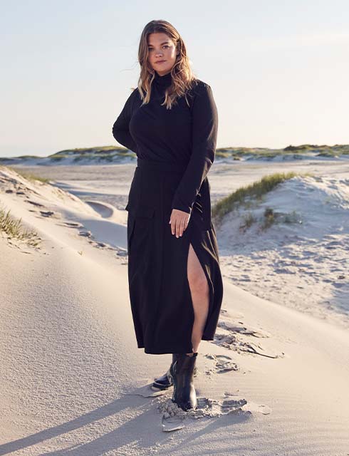 The model wears a blue dress and stands in a dune landscape.

