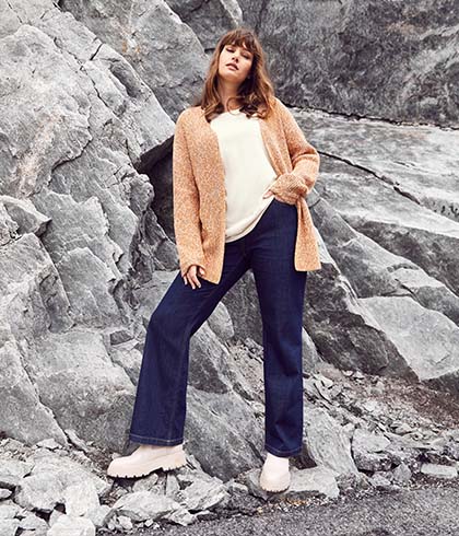 Woman in an orange cardigan and blue jeans stands in front of a rock face 