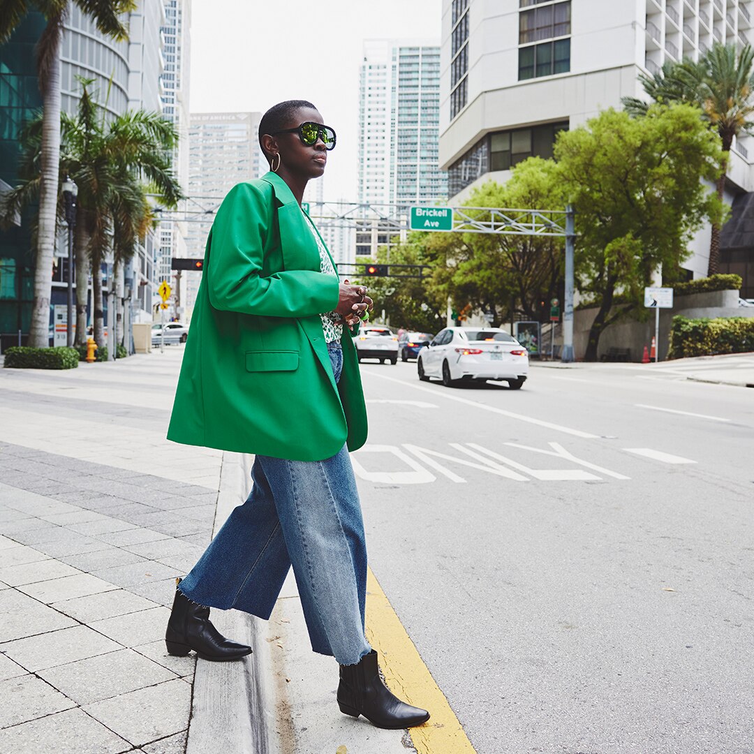 Woman with a long green blazer and wide jeans is walking across the street