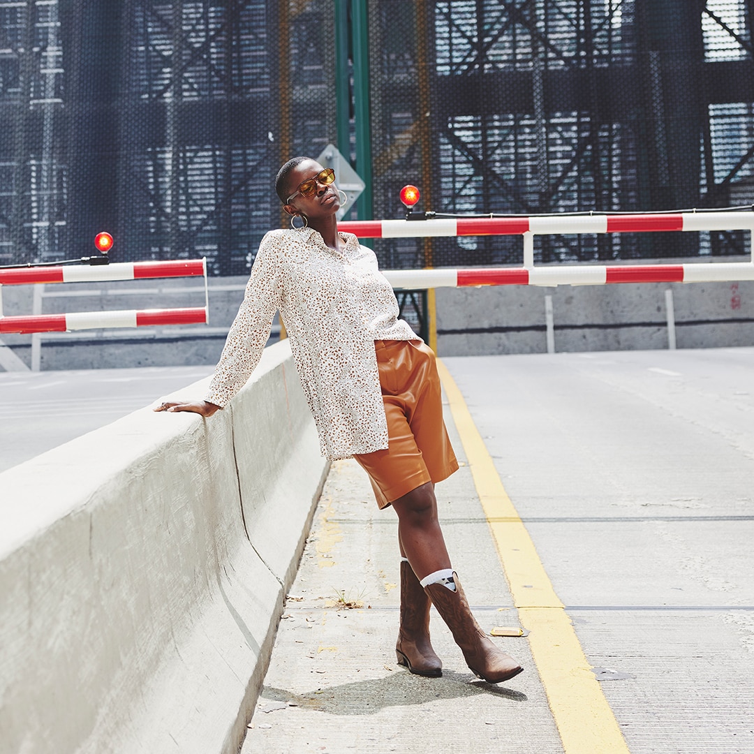 Model with a long blouse, a short leather pants and cowboy boots is leaning against a barrier