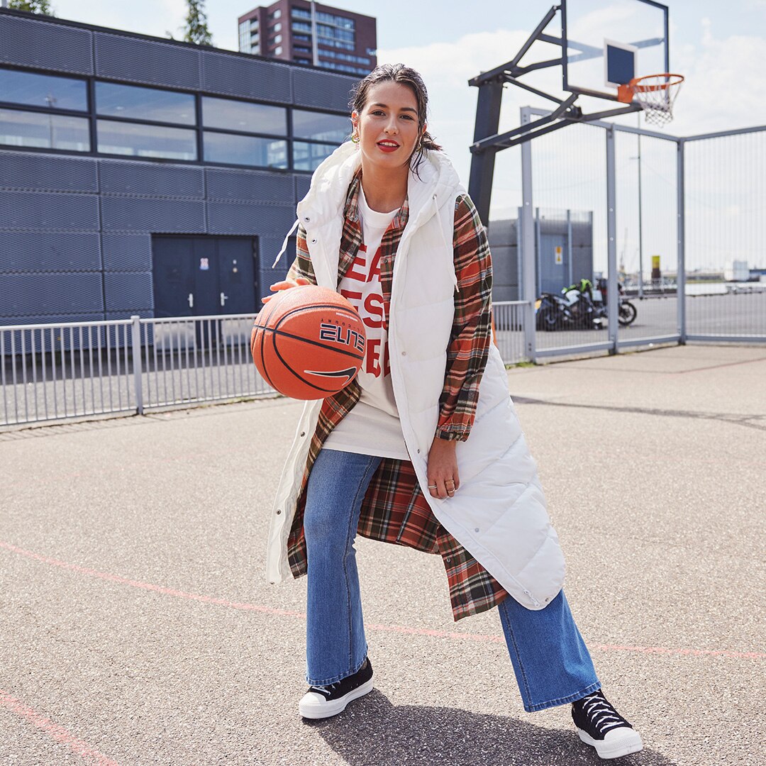 Model is standing on a basket ball field wearing a white long vest and holding a baskett ball in her hand