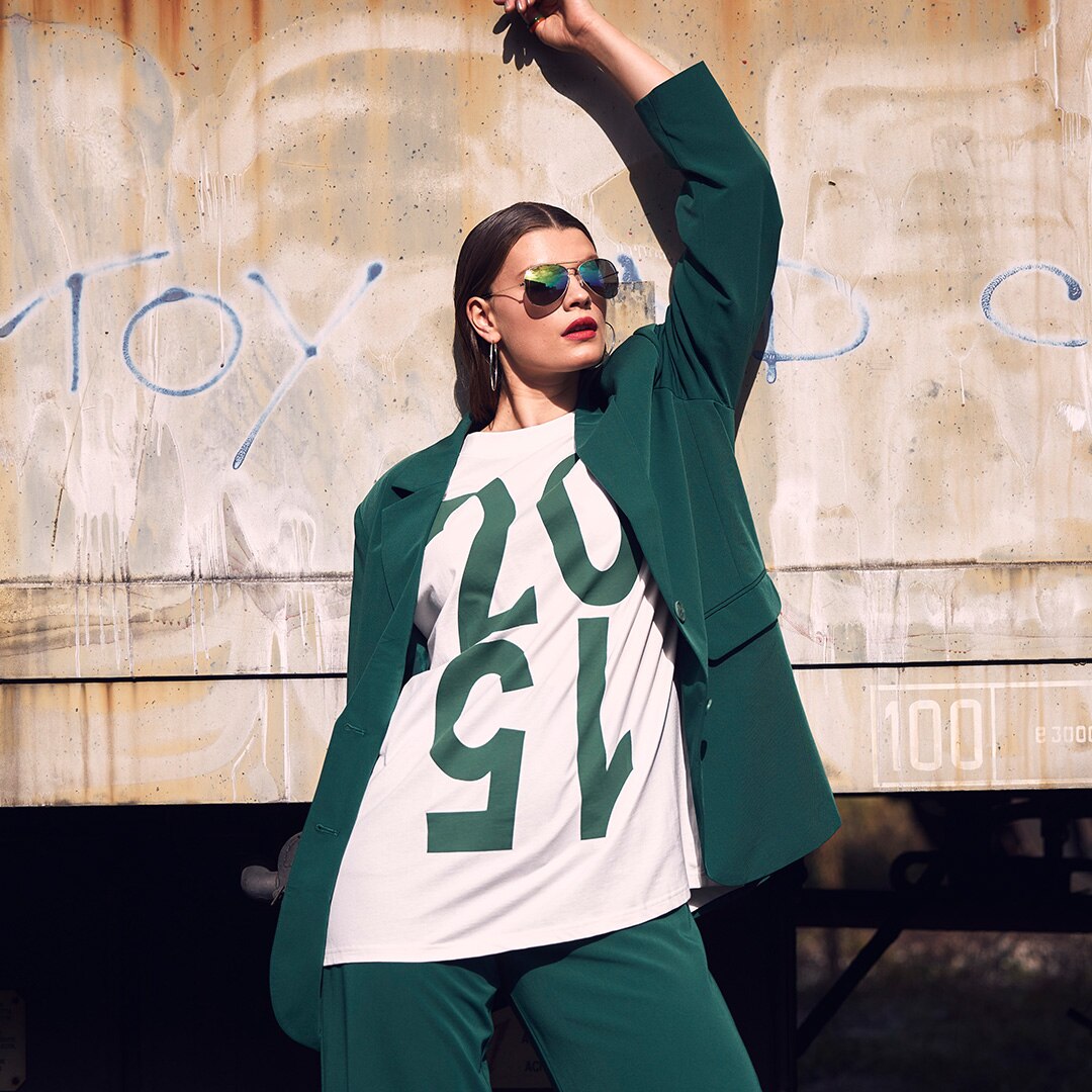 Model is wearing a white t-shirt and a green blazer while standing in front of a wall