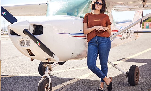 Model leaning against an airplane wearing jeans and sunglasses