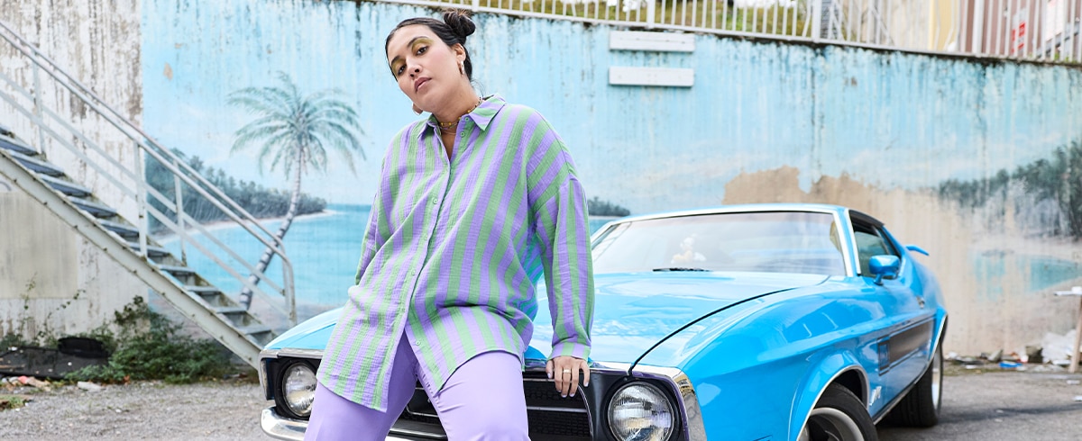 The model is wearing a purple and green striped blouse and matching tight-fitting purple trousers.