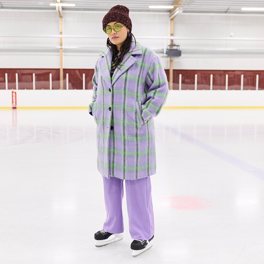 The model is standing on the ice wearing ice skates and a green and purple checked coat.