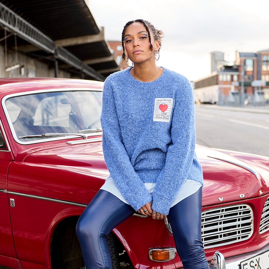 The model is wearing a green woollen jumper and purple trousers.