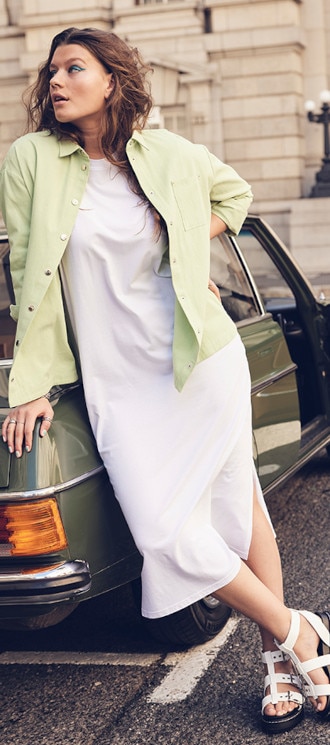 Woman in a white dress and green shirt stands leaning against a car in front of a building.