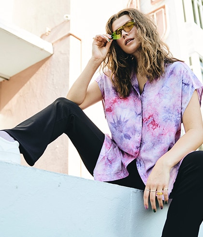 Model sitzt auf einer Mauer mit Sonnenbrille und lila Bluse.