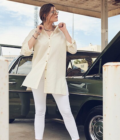 Woman poses in front of a green vintage car wearing a long beige blouse and white jeans.