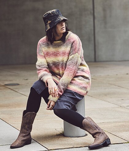 Model sitting on a pillar with a sweater and a hat on. 