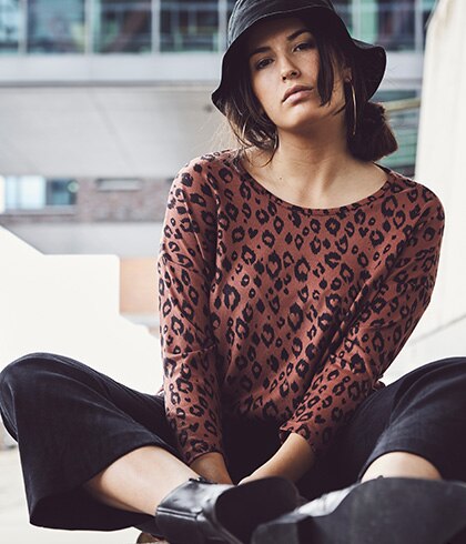Woman with a black hat on and wearing a leoprint shirt is sitting in front of a building.