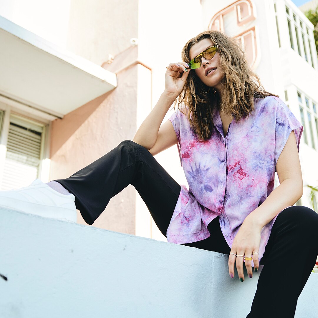 Model sitzt auf einer Mauer mit Sonnenbrille und lila Bluse.