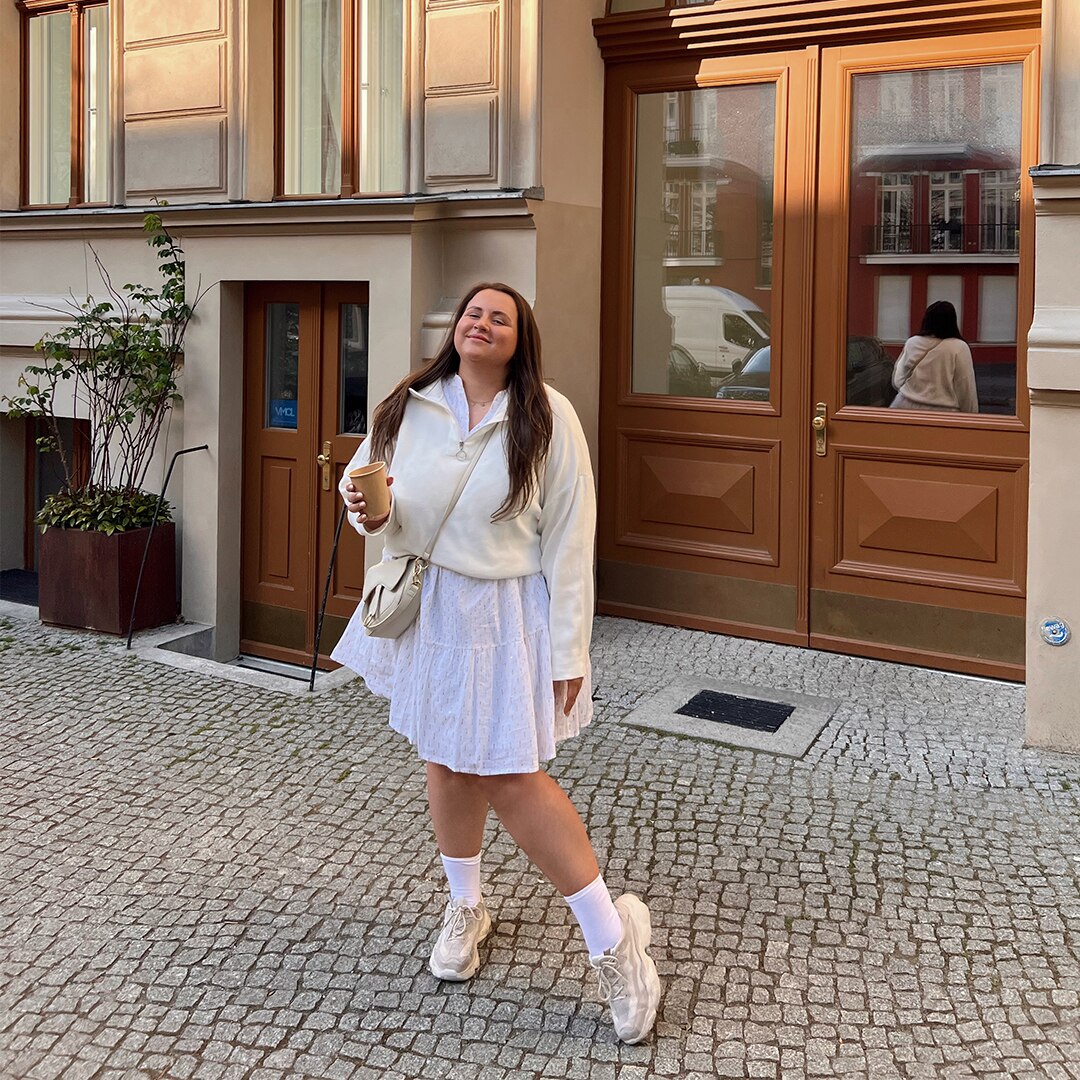 Woman holding coffee mug, wearing a white dress, jumper and sneakers poses in front of a brown entrance door.