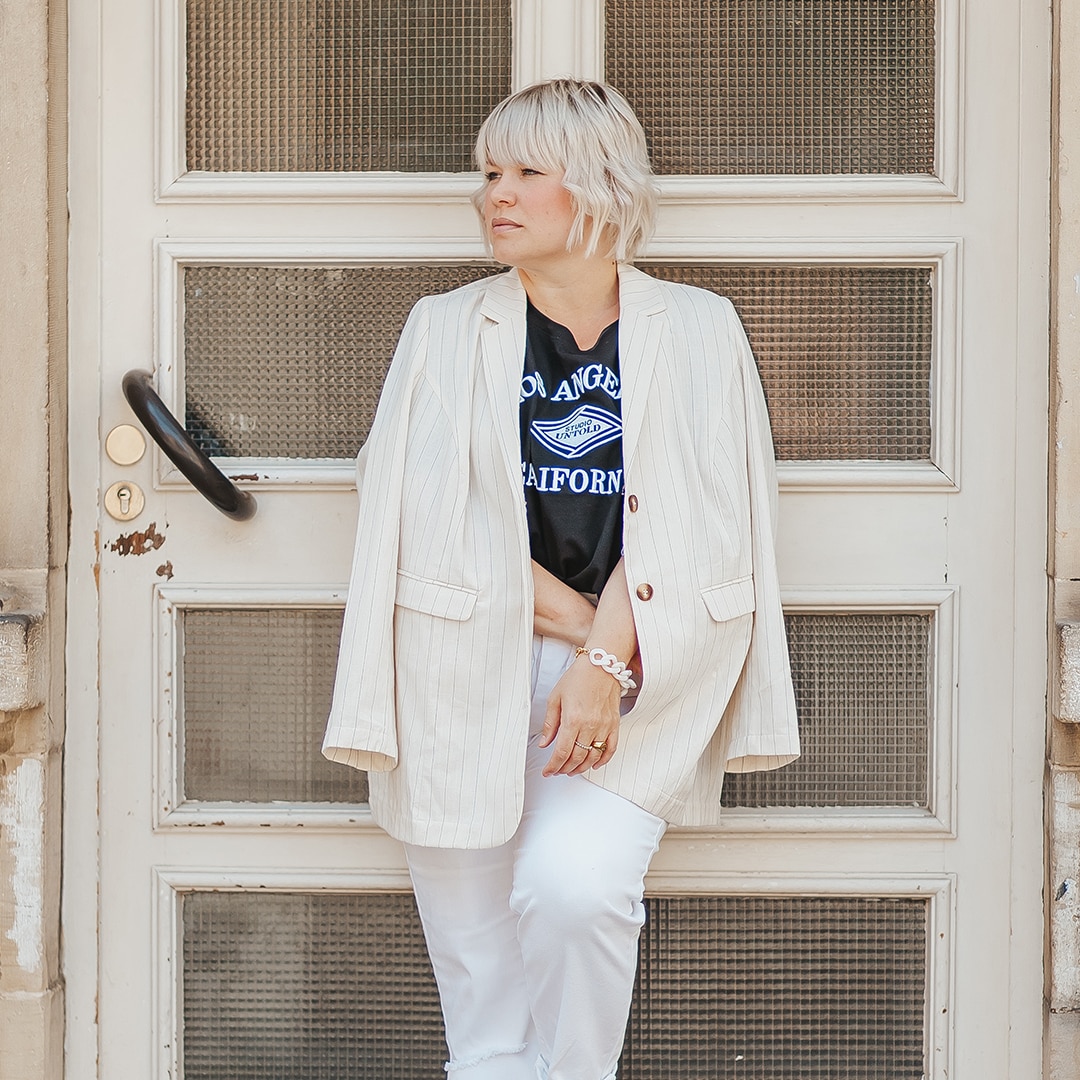 Woman with blonde hair leaning against a big door, wearing a beige longblazer, white jeans and a black t-shirt.