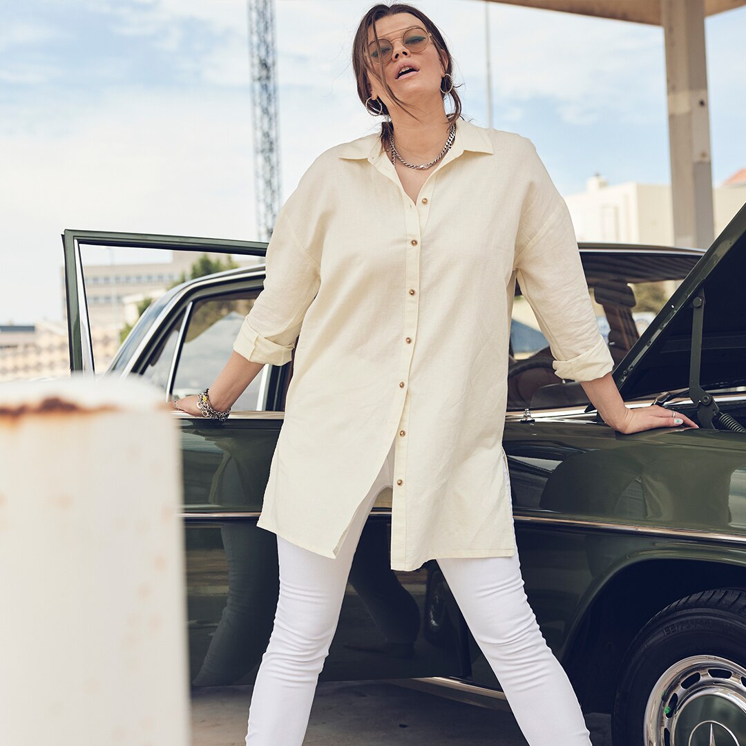 Woman wearing a beige shirt and white jeans poses in front of a green vintage car.