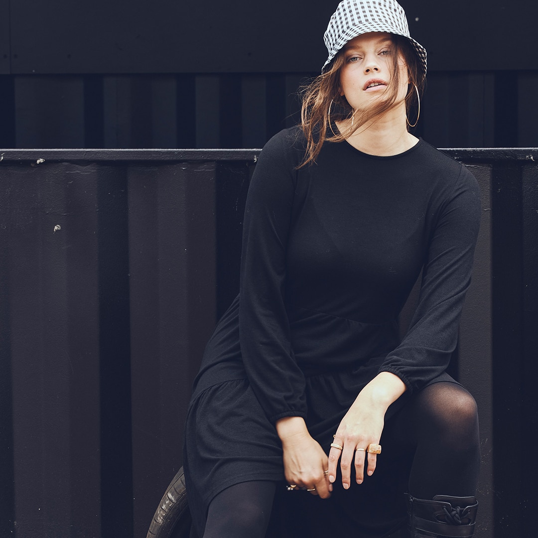 Model with black clothes and a light hat on is posing in front of a black wall