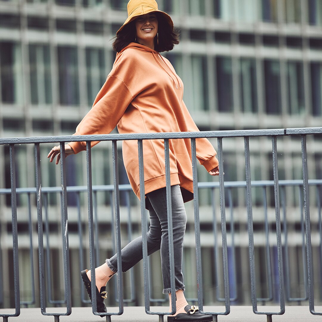 Model posiert auf einer Brücke in einer orangen Jacke und mit Hut auf