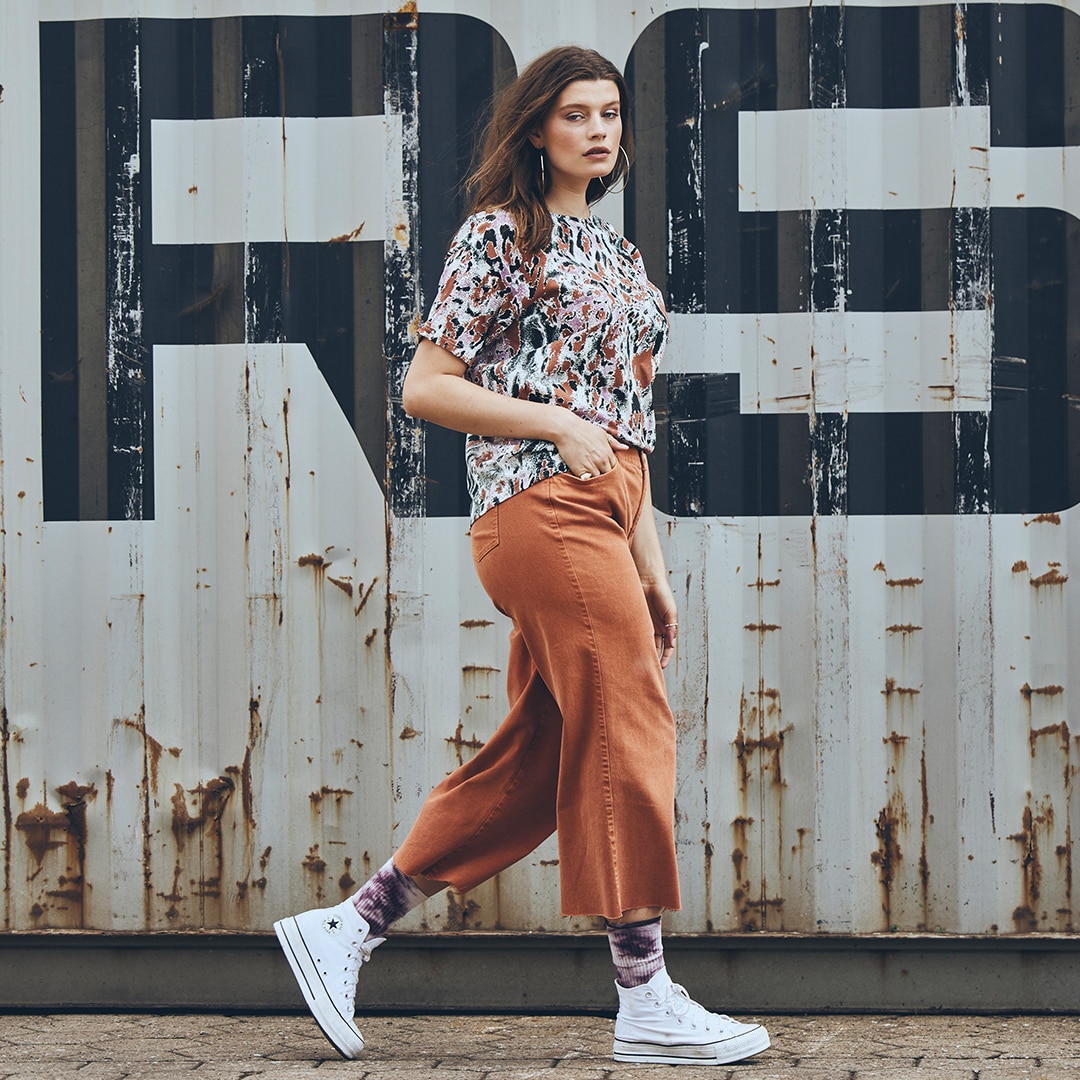 Model mit oranger Culotte Hose und weißen Sneakern vor einem Container