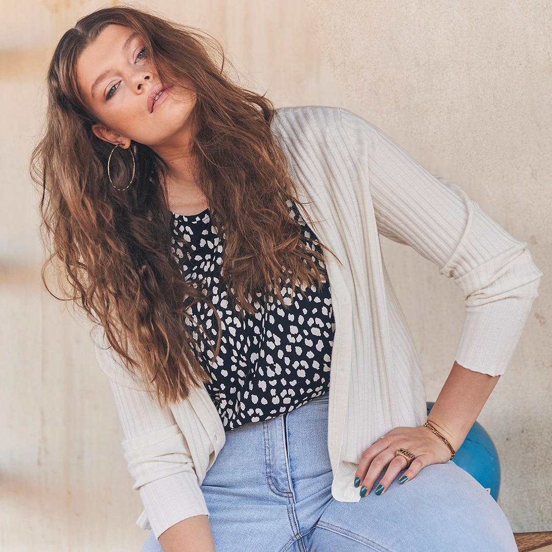 Woman with brown curly hair, wearing a ripped cardigan, jeans and an animal print blouse.