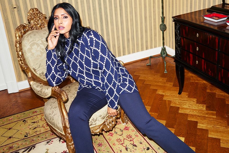 The model sits on a suitcase and wears a blue long blouse with white jeans