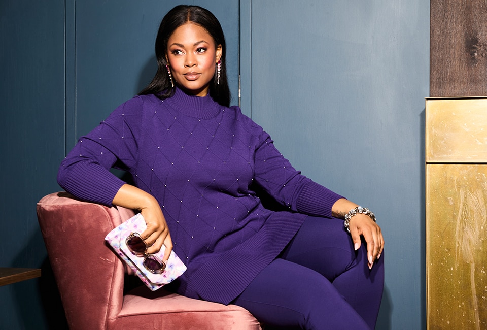 The model is sitting on a couch wearing a purple sweater.