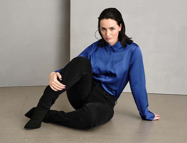 The model leans sitting on the floor and wears a blue blouse 