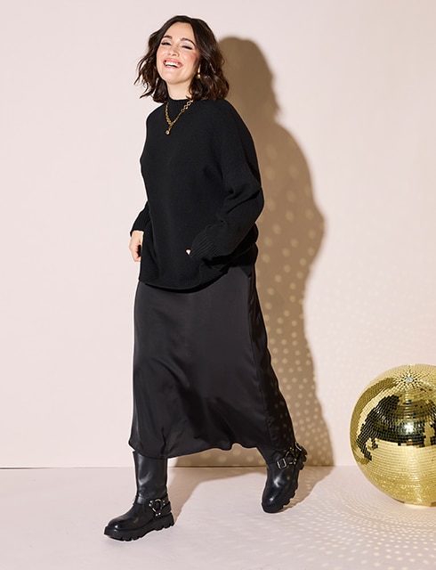 The model poses next to a golden disco ball in a black satin skirt and a black turtleneck sweater