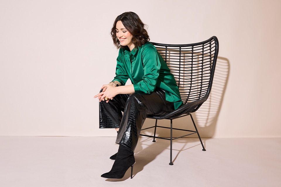 The model poses sitting on a chair and wears black imitation leather pants and a green blouse