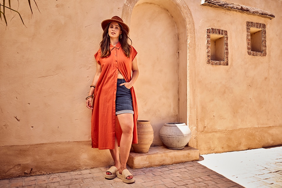 The model is wearing a long, orange blouse and short jeans.