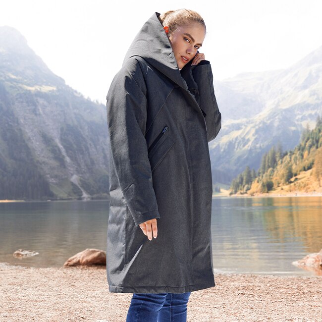 The woman wears a grey jacket in front of the lake 