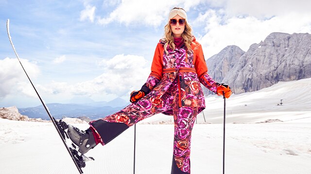 Blond model in orange and pink skiwear poses with ski equipment