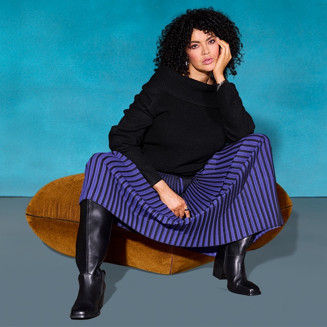 The model sits on a chair and wears a purple pleated skirt and a black sweater.