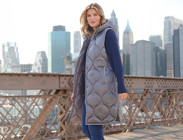 The model poses in a blue sweater with a long vest on a bridge in New York