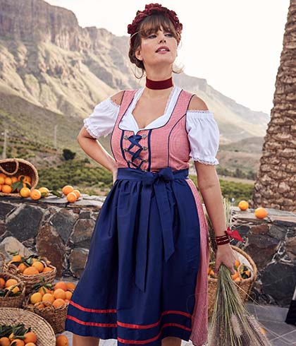 The model wears a dirndl while posing in front of baskets filled with oranges
