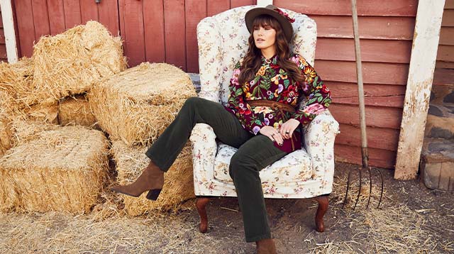 The model sits in an armchair in front of the barn and wears a colorful blouse 