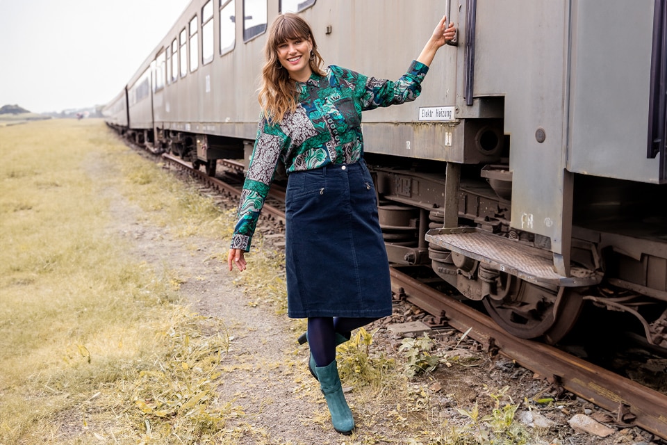 The model poses at a train in a patterned blouse and a corduroy skirt  
