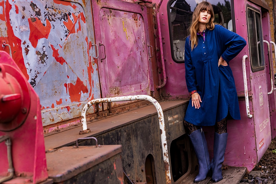 The model poses by a pink wagon and wears a blue corduroy dress