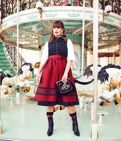 Woman in a dark dirndl with red apron poses in front of a carousel
