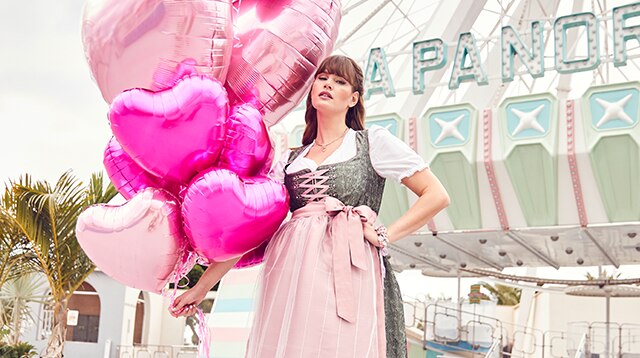 Woman in dirndl standing in front of Ferris wheel with pink balloons