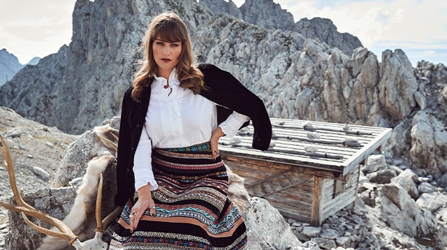 Woman with patterned skirt, white blouse and velvet jacket sitting on a rock.