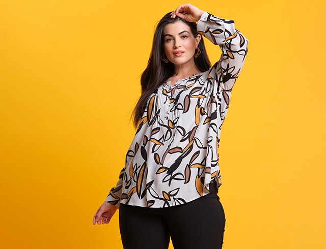 Dark-haired woman in front of a yellow wall wears a colourful blouse and poses