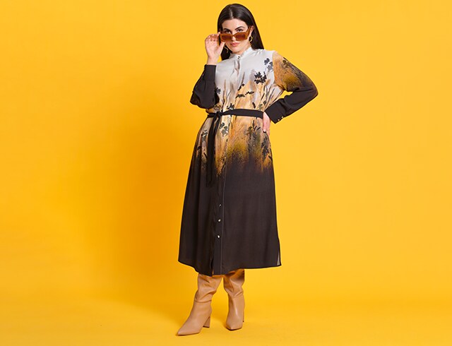 Dark-haired woman posing in front of yellow wall in a colourful dress with sunglasses