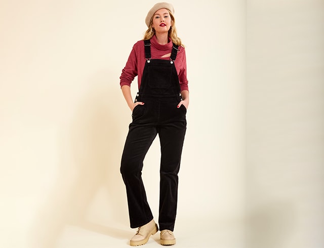 A blonde woman standing in front of a beige wall wears dungarees, a roll-neck jumper and a cap