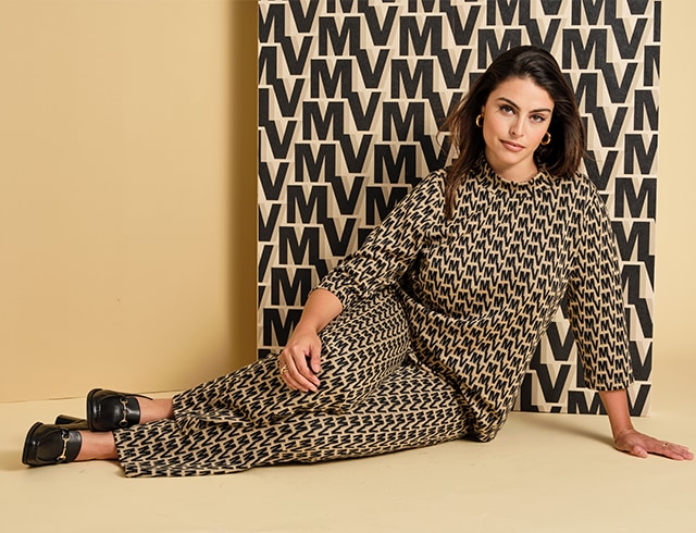 Dark-haired woman sits in front of patterned wallpaper and wears a top and trousers with the same pattern