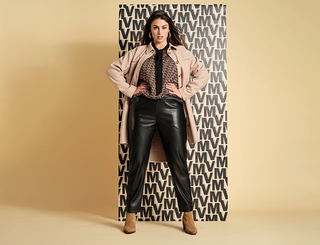 Dark-haired woman in front of a patterned wall wearing a colourful blouse, leather trousers and a coat