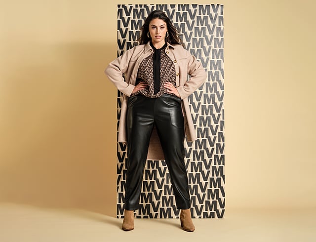 Dark-haired woman in front of a patterned wall wearing a colourful blouse, leather trousers and a coat