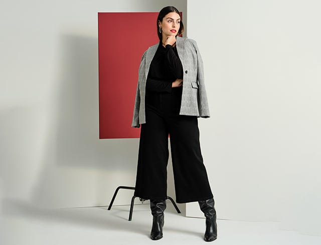 Dark-haired model posing in front of a red wall in a black outfit with a grey jacket 